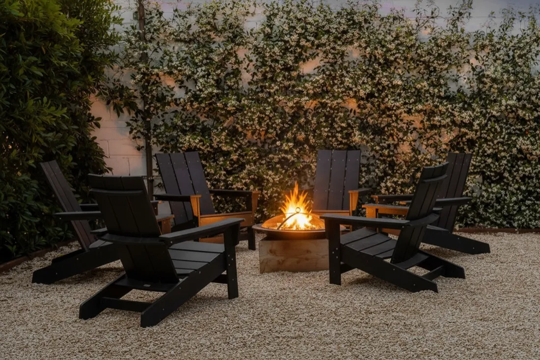 Lit fire pit ringed by Adirondack chairs on a gravel patio at dusk